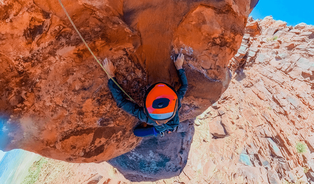 Overhead view of a climber in an orange Petzl helmet gripping a red rock overhang with a canyon drop below.