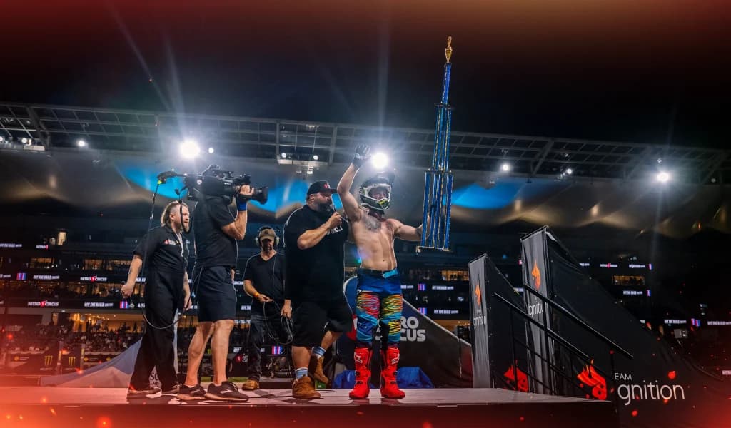 A motocross rider raises a tall trophy on a stadium stage while crew members and a camera operator stand nearby.