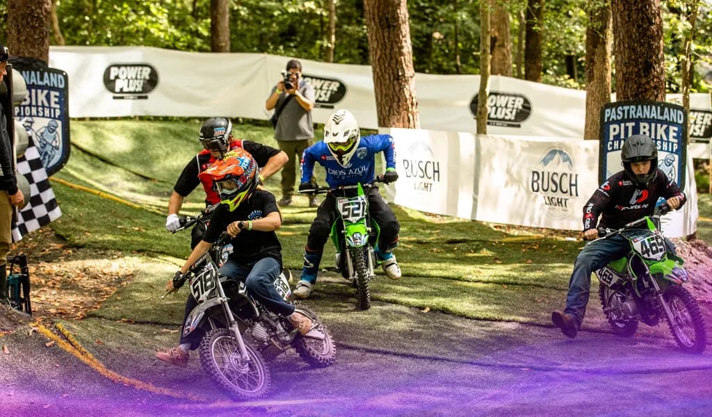 Group of pit bike riders at Pastranaland accelerating from the start of a race on a shaded outdoor track.