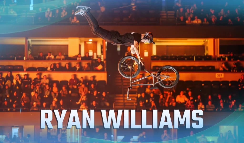 BMX rider airborne above an arena crowd while the bike rotates upside down beneath him during a stunt.