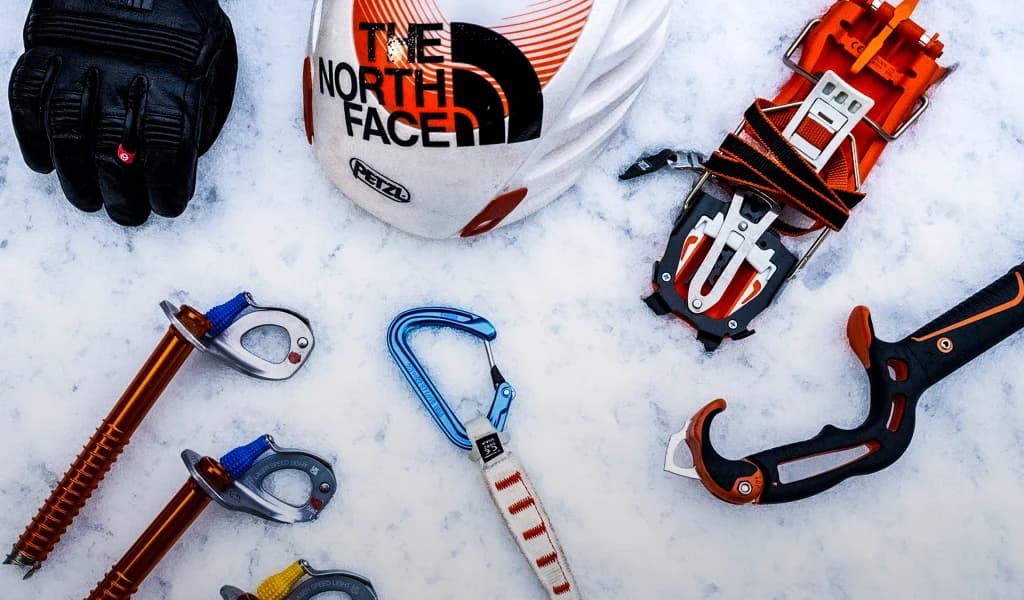 Ice climbing gear arranged on snow, including a helmet, crampons, ice screws, carabiners, gloves, and an ice tool.