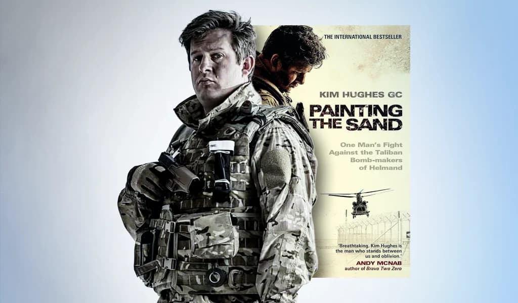 Portrait of Kim Hughes in military gear beside his book “Painting in the Sand” about bomb disposal in Afghanistan.