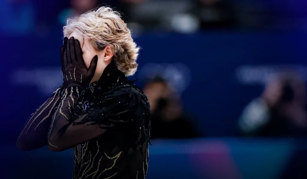 Ilia Malinin covering his face with gloved hands after a figure skating performance.