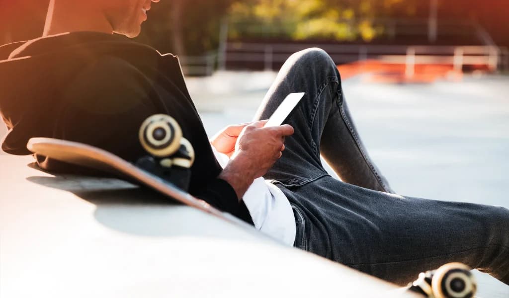 Skateboarder sitting at a skatepark while using a smartphone, with a skateboard resting beside them.
