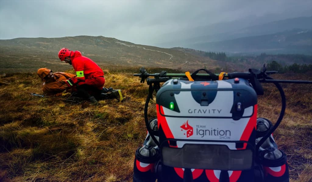Rescue worker assisting an injured person on a hillside, with a Gravity Industries jet suit in the foreground displaying the Team Ignition logo.