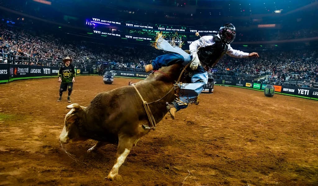 Bull rider being thrown from a brown bull at a PBR arena event with a bullfighter visible in background.