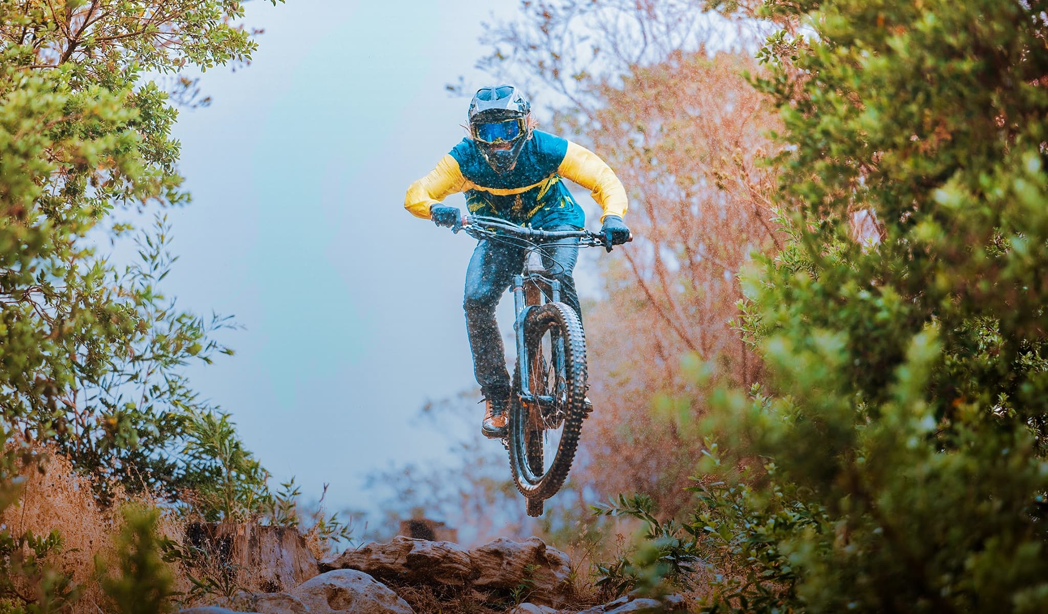 Mountain biker in yellow and blue gear launching off a rock drop through trees with mist in the background.
