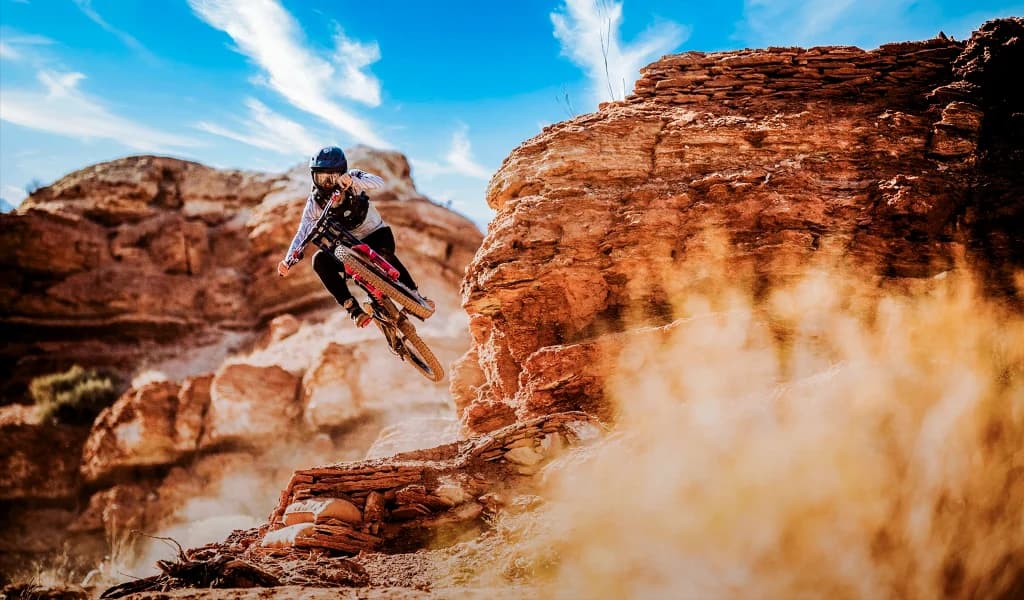 Mountain biker launching off a red rock ledge in a dust cloud under a blue sky in a desert landscape.