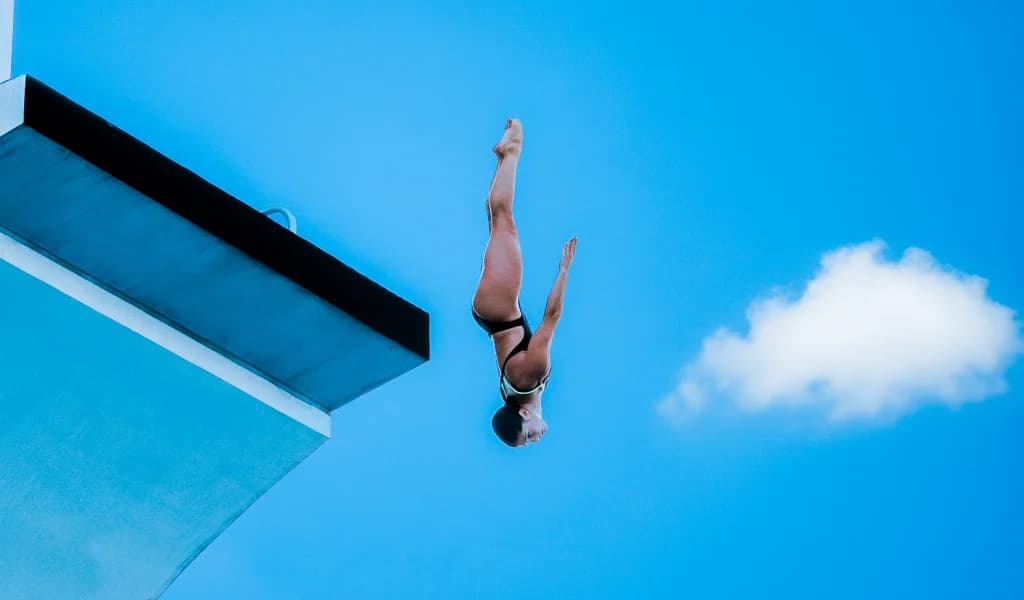 Morgane Herculano in a black swimsuit falling headfirst from a high diving platform against a blue sky.