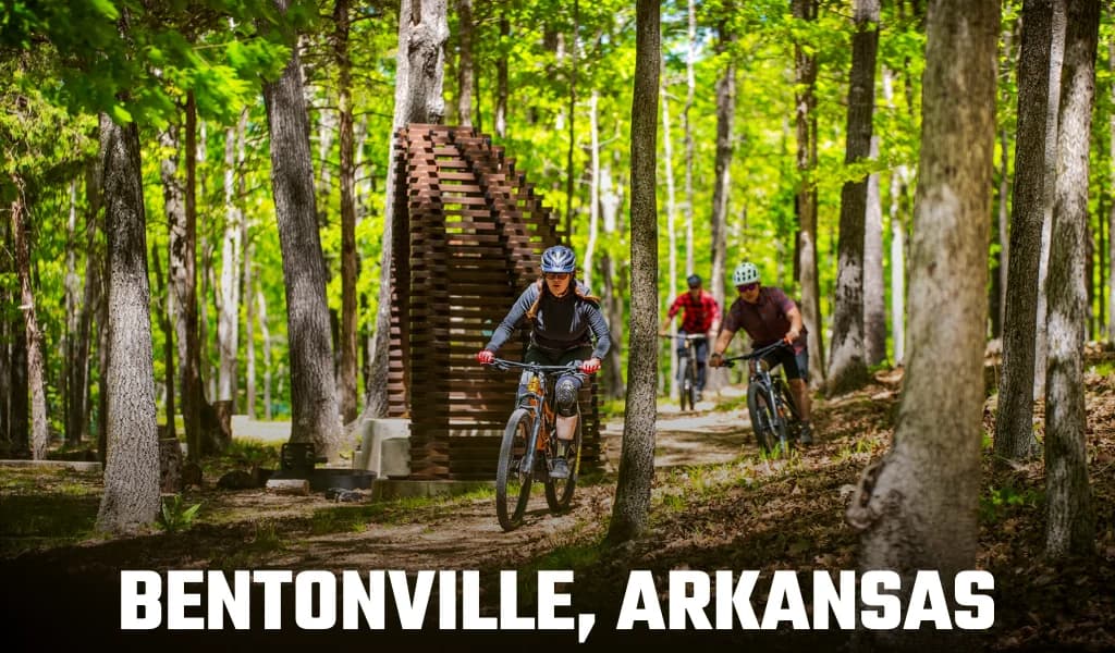 Text reading Bentonville, Arkansas over three mountain bikers riding a forest trail past a wooden art installation.