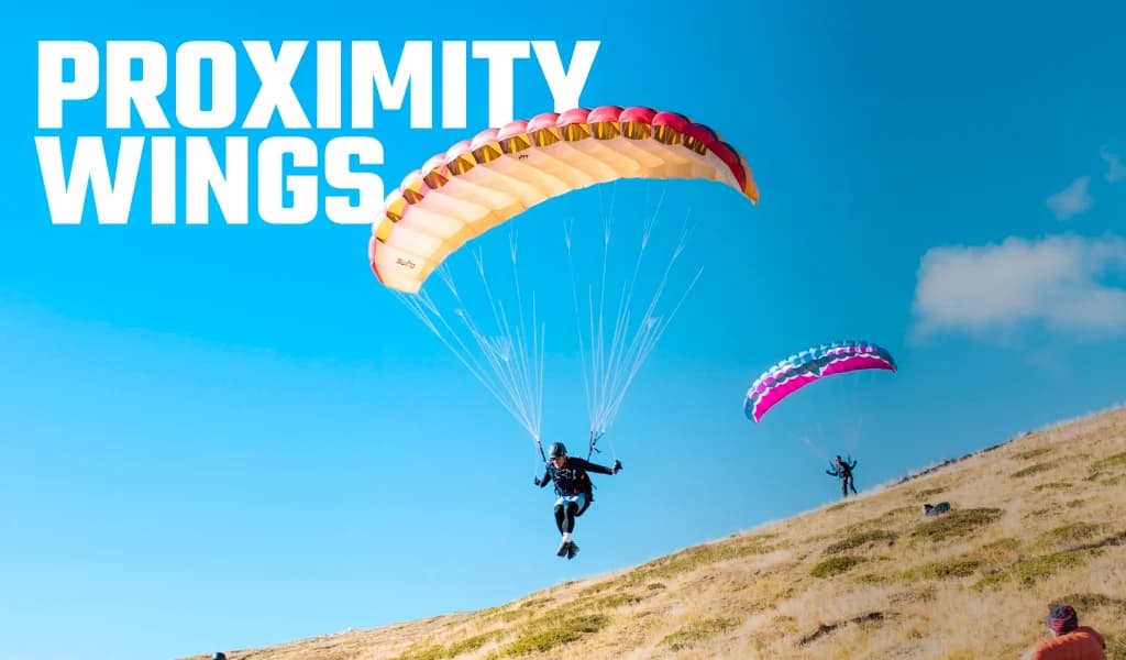 Text reading Proximity Wings, over two pilots landing speed wings on a grassy hillside.