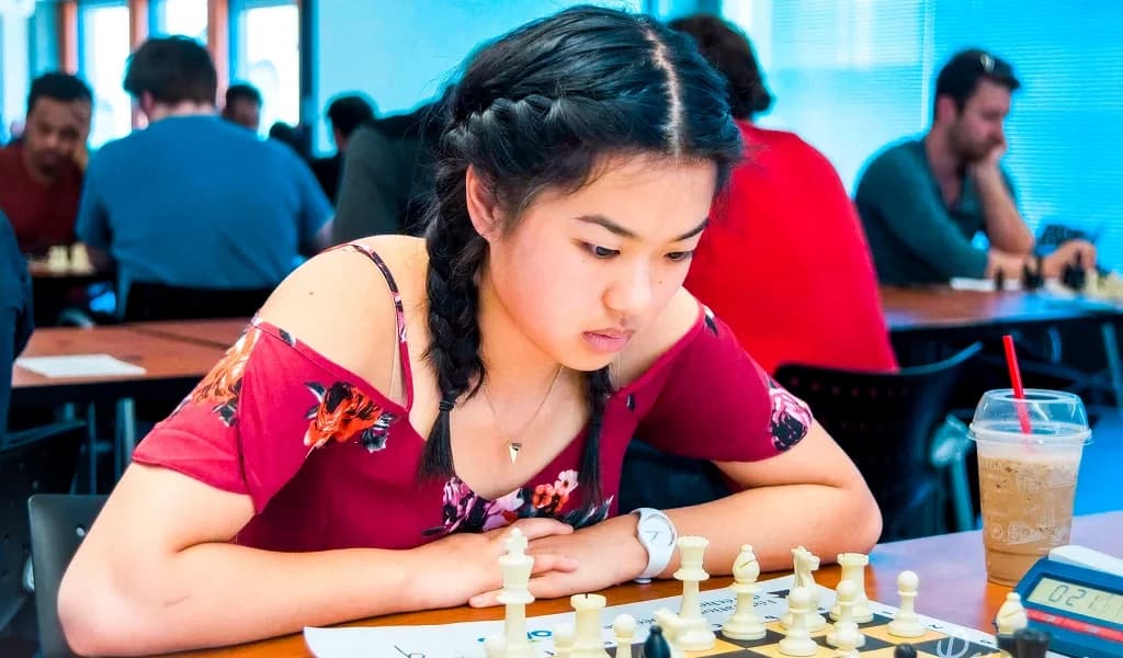 Nemo Zhou studies a chessboard intently at a tournament table, iced coffee and chess clock beside her.