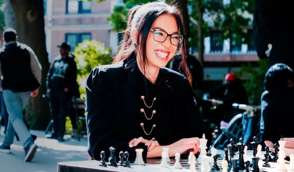 Nemo Zhou smiles while playing chess outdoors at a park table, wearing glasses.