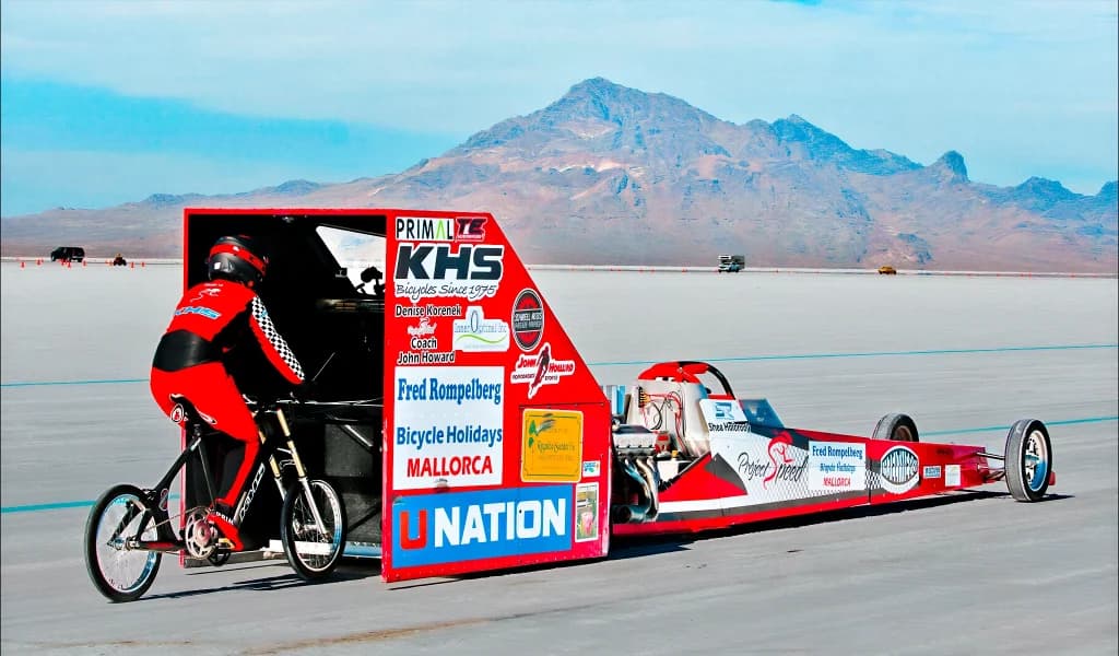 Cyclist riding behind a streamlined dragster on a salt flat during a speed record attempt.