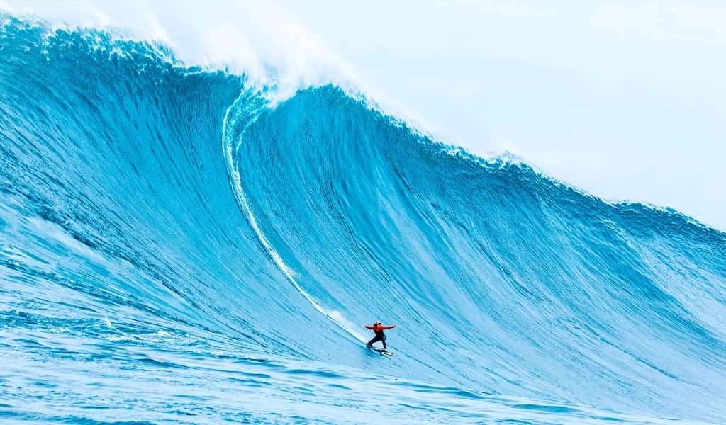 A surfer rides a massive blue wave with arms outstretched, dwarfed by the wall of water.