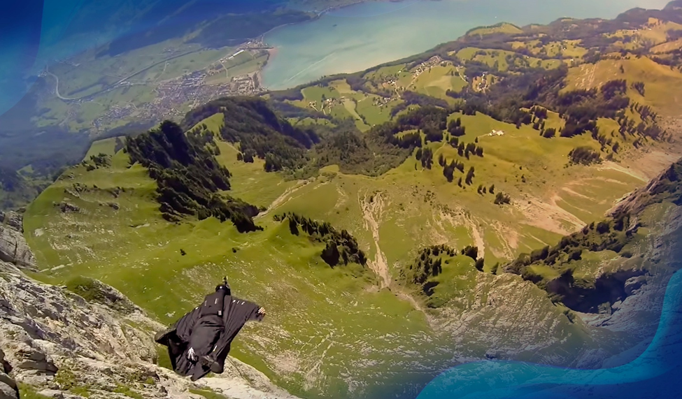 Jeb Corliss gliding in a wingsuit over a vast, mountainous landscape in Switzerland, with a view of the valley below.