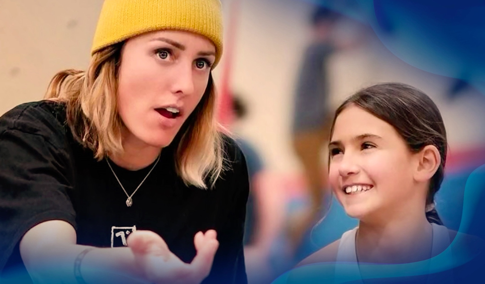 Alex Johnson wears a yellow beanie, explaining something passionately to a young girl who listens attentively, both smiling in a casual indoor setting.