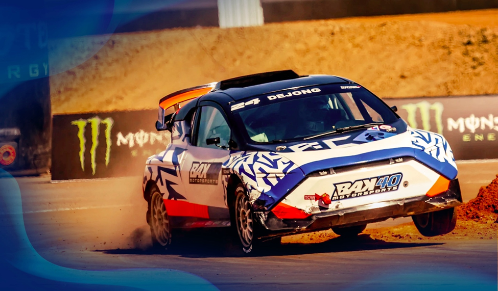 A blue and white race car driven by Dejong skids on a dirt track at a Nitro Crosse event. Monster Energy signage appears in the background.