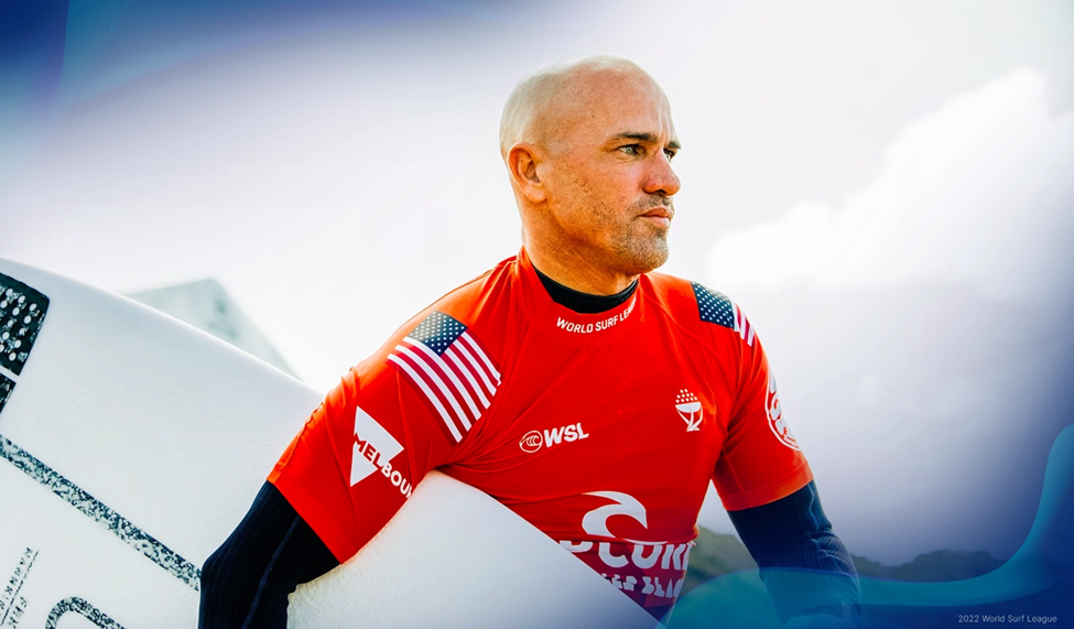 Kelly Slater in a red competition jersey holding a surfboard, looking focused with a blurred background.