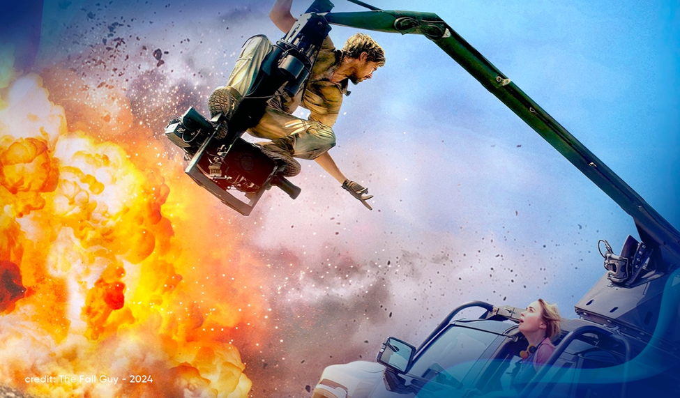 Ryan Gosling swings from a crane while shooting an action scene with an explosion with fire and a shocked woman played by Emily Blunt in a nearby vehicle.