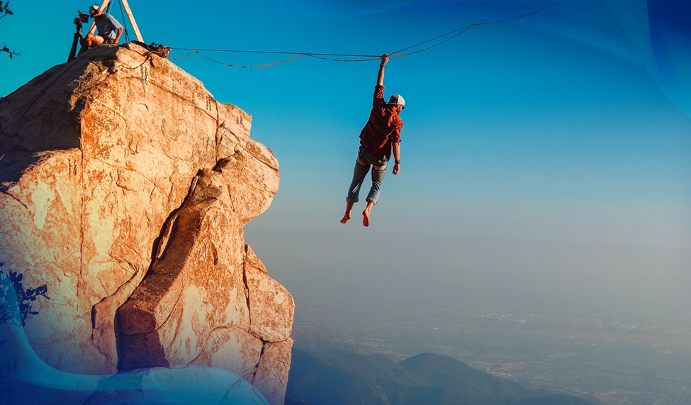 Ryan Robinson hangs by one hand from a highline above a cliff, with a vast valley and clear blue sky in the background.