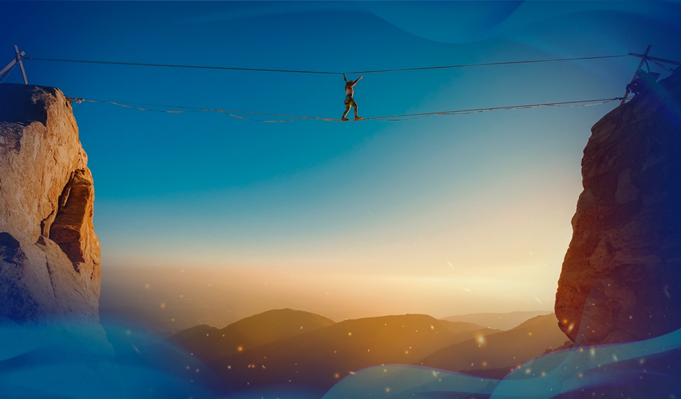 Leticia Bufoni walks across a slack rope suspended hundreds of feet in the air between two cliff faces. A blue sky and a mountain range make up the backdrop.