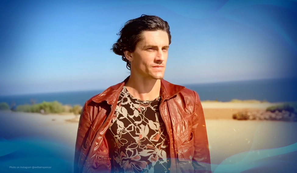 William Spencer in a leather jacket, standing near the ocean under a clear blue sky.