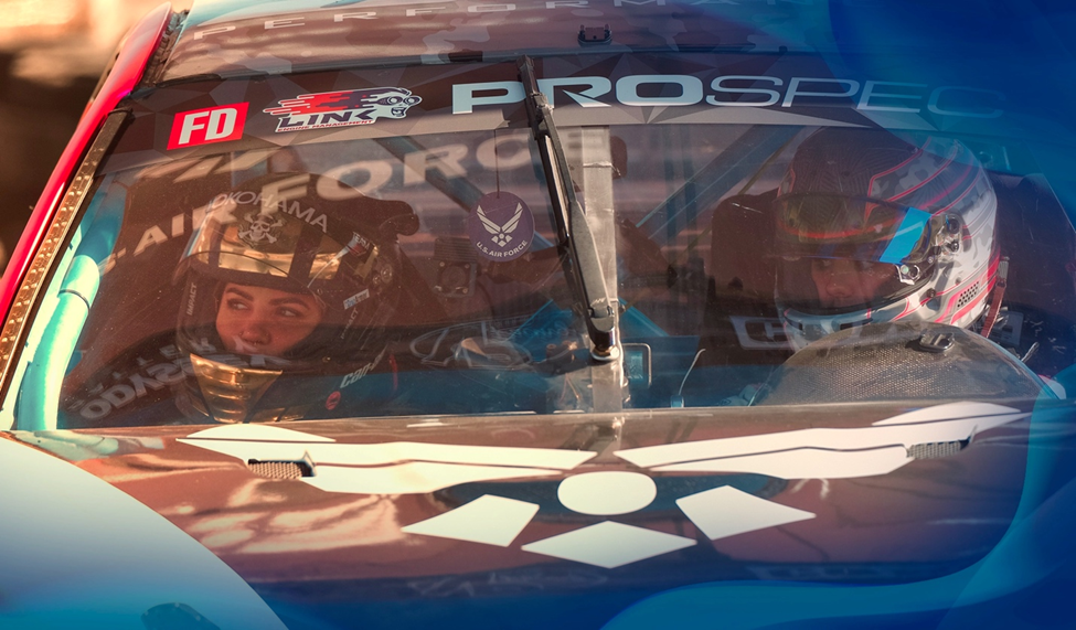 Amanda Sorenson seated in a race car wearing a helmet, her focused expression visible through the windshield with logos and decals in the foreground.