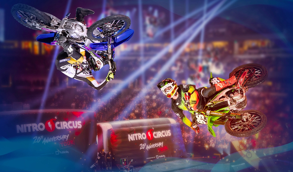 Two motocross riders performing high-flying tricks on dirt bikes in an arena with spotlights and a Nitro Circus 20th Anniversary Tour logo in the background.