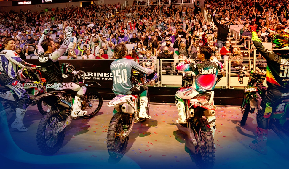 Nitro Circus team riders seated on motorcycles celebrating in front of a cheering crowd inside a brightly lit stadium.