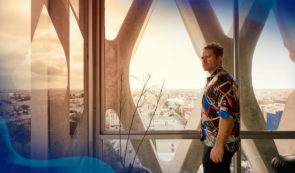 Daniel Cates in a vibrant patterned shirt stands beside a floor-to-ceiling window overlooking a sprawling city, framed by curved architectural beams.