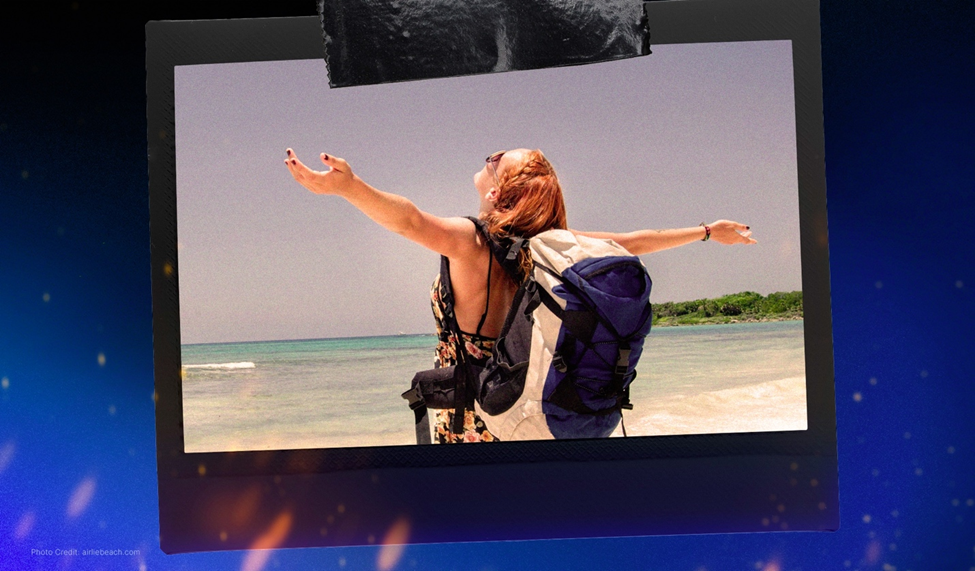 A woman with a backpack standing on a sandy beach, arms outstretched toward the ocean, enjoying the view under a clear sky.